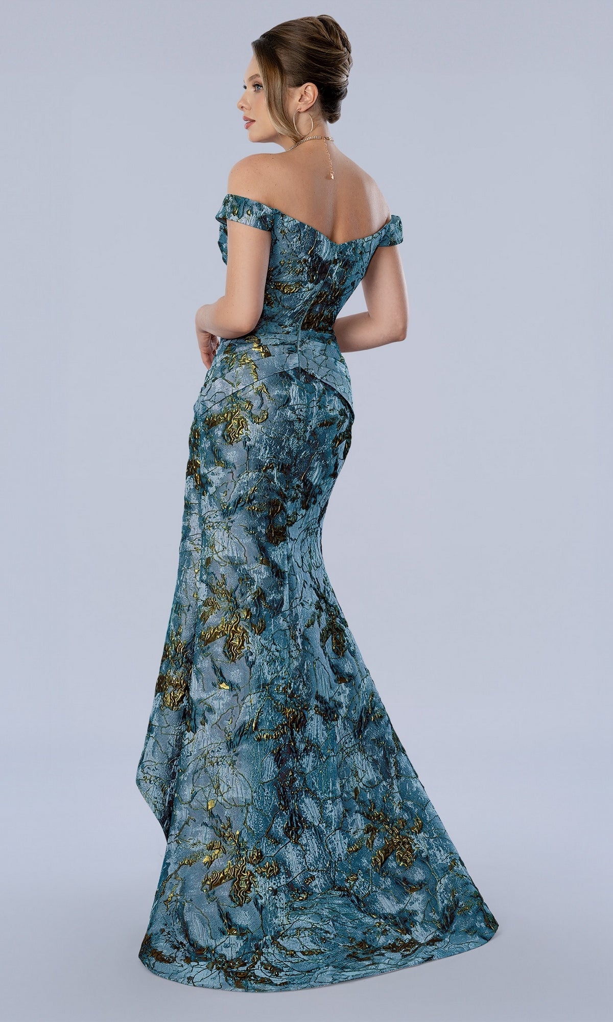 A woman wears a Stella Couture 26361 green long formal dress, an off-the-shoulder, floor-length mermaid gown with a metallic gold and blue textured pattern.
